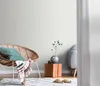 A cozy living room corner with a wicker chair, a small wooden side table with a vase and dried flowers, and a light blue armchair in the background. Produktbild-Vorschau