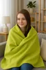 A young woman is sitting on a sofa, wrapped in a lime green blanket. Produktbild-Vorschau