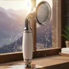 A retro-style shower head with a ceramic handle is shown on a wooden surface, with water droplets falling and a blurred background of mountains and a window. Podgląd zdjęcia produktu