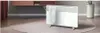 A white electric heater with a towel draped over it, placed on a rug in a room with a plant and a wall-mounted shelf. Produktbild-Vorschau