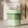A collapsible washing machine with a drain hose attached, placed on a tiled floor in front of a wooden cabinet. Náhled obrázku produktu