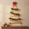 A wall-mounted Christmas tree decoration made of wooden shelves, adorned with pine branches, berries, pinecones, ornaments, and LED lights, topped with a red bow. Several gift-wrapped boxes are placed on a wooden dresser below. Náhled obrázku produktu