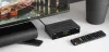 A black DVB-T2 tuner and remote control are placed on a white surface next to a television. Náhled obrázku produktu