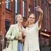 Two young women are taking a selfie on a city street with brick buildings in the background. Produktbild-Vorschau
