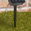 A close-up view of a solar-powered garden light stake inserted into artificial grass, with a brick wall in the background. Produktbild-Vorschau