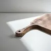 A wooden cutting board with a handle is placed on a light-colored surface with shadows. Anteprima dell’immagine prodotto