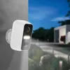 A white security camera mounted on a gray wall at dusk, with a blurred background showing a modern house and garden. Náhľad obrázku produktu