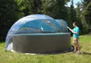 A man is standing next to a round pool with a transparent dome cover in a grassy outdoor area with trees in the background. Produktbild-Vorschau