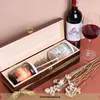 A wooden wine box containing two jars and a honey dipper, with a bottle of red wine and a glass of wine next to it, and dried flowers in the foreground. Produktbild-Vorschau
