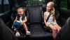 Two young girls are sitting in car seats in the back seat of a car. Voir les images du produit