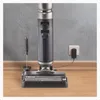 A cordless floor cleaner is shown plugged into a wall outlet for charging, with a cleaning tool resting beside it. Produktbild-Vorschau