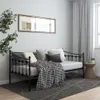 A metal daybed with pillows and a blanket is shown in a room with a plant and curtains. Náhled obrázku produktu