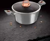A pot with a glass lid and copper accents sits on a dark, textured surface with scattered copper-colored pieces. Náhled obrázku produktu