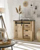 A rustic wooden sideboard with sliding doors and drawers is displayed in a well-lit room with a white brick wall background. Decorative items like dried pampas grass, vases, and framed pictures are arranged on and around the sideboard, creating a cozy and  Produktbild-Vorschau