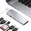 A silver USB-C hub is shown connected to a laptop, with various accessories like USB cables and memory cards laid out around it. Produktbild-Vorschau
