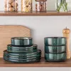 A set of dark green ceramic plates and bowls are neatly stacked on a wooden shelf, with a wooden cutting board and a pepper mill visible in the background. Voir les images du produit