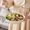 Eine Frau in einem beigen Kleid hält eine weiße Salatschüssel mit Salat und isst mit einer Gabel. Náhled obrázku produktu
