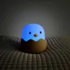 A cute chick-shaped night light is glowing blue inside a brown eggshell, resting on a textured surface. Produktbild-Vorschau