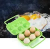 A green egg carton is shown in the foreground, with eggs inside and some empty compartments. In the background, fried eggs are cooking on a grill over aluminum foil, with smoke rising. Produktbild-Vorschau