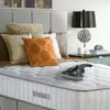 A hand-held vacuum cleaner is placed on a mattress next to several pillows, with a bedside table and lamp in the background. Produktbild-Vorschau