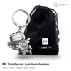 A silver angel keychain with a clover is displayed next to a black velvet pouch, with brand logos and text visible. Produktbild-Vorschau