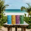 Four tiki mugs are displayed on a wooden table in front of a tropical beach scene. Podgląd zdjęcia produktu