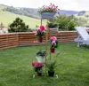A gold metal plant stand with various potted plants on a green lawn, with a wooden fence and rolling hills in the background. Náhľad obrázku produktu