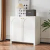 A white cabinet with arched doors and gold handles, placed in a room with wooden flooring and a brown curtain. Produktbild-Vorschau