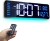 A hand holding a remote control in front of a digital wall clock displaying the time, date, and temperature. Produktbild-Vorschau