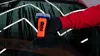 A person wearing a red sleeve and black glove is holding an orange product with a blue cap on a wet car window. Produktbild-Vorschau
