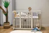 A baby is sitting in a white crib next to a potted plant. A stuffed bear is lying on the floor in front of the crib. Produktbild-Vorschau