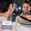 A man is brushing his teeth in front of a mirror, holding an electric toothbrush. Produktbild-Vorschau