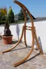 A wooden hammock stand is displayed outdoors on a stone patio, with a palm tree in a pot to the left and a white wall and houses in the background. Produktbild-Vorschau