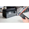 A black Krüger&Matz KM0816 audio device is shown on a table, with a hand holding a remote control pointing towards it. Several vinyl records are leaning against another device in the background. Produktbild-Vorschau