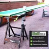 A long green plank is supported by two black metal sawhorse-style stands. A can of paint sits on the plank. The scene appears to be outdoors on a paved surface. Produktbild-Vorschau