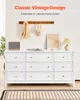 A white dresser with nine drawers is shown in a room setting, with decorative items on top and a coat rack on the wall. Náhled obrázku produktu