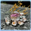 A collection of clear resin cubes and a sphere containing preserved flowers, arranged on a dark, textured surface with dried yellow flowers in the background. Voir les images du produit