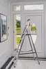 A silver multi-purpose ladder is positioned in a hallway in front of a white door with a large oval window. The ladder has yellow accents on its steps and feet. The floor is tiled with a black and white striped pattern. Náhled obrázku produktu