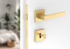 A close-up view of a gold-colored door handle set, including the handle, lock mechanism, and keyhole plate, mounted on a white door. The background shows a blurred interior of a bedroom. Produktbild-Vorschau