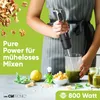A person is holding a black and silver immersion blender over a glass container with green liquid, surrounded by ingredients like walnuts, lemon, basil, and cheese. Voir les images du produit