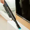 A hand is holding a black cordless vacuum cleaner and cleaning the edge of a white surface next to a television. Náhled obrázku produktu