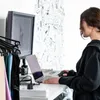 A woman is working at a desk with a laptop and monitor, with fabric samples hanging on a rack to the left. Podgląd zdjęcia produktu