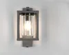 A modern outdoor wall lamp with a glass cylinder and a gray metal frame is mounted on a white wall. Anteprima dell’immagine prodotto