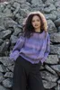 A woman stands in front of a stone wall, wearing a knitted sweater. Produktbild-Vorschau