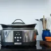 A Crockpot slow cooker is shown on a kitchen counter, with cooking utensils and carrots in the background. Produktbild-Vorschau