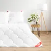 A white duvet and pillows are neatly arranged on a bed, with a small side table holding a vase of flowers and decorative items next to a floor lamp. Produktbild-Vorschau