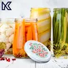 A collection of jars filled with preserved vegetables, including cauliflower, carrots, and green beans, arranged on a white wooden surface. A decorative lid with a floral pattern is placed in the foreground. Anteprima dell’immagine prodotto