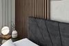 A close-up view of a bed with a dark upholstered headboard and a wooden slatted wall behind it. A bedside table with a lamp and a vase is also visible. Produktbild-Vorschau