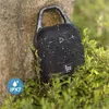 A black portable Bluetooth speaker with a carabiner hook is attached to a tree trunk, surrounded by green foliage. Water droplets are visible on the speaker. Voir les images du produit