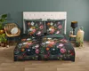 A bedroom scene with a bed dressed in floral duvet cover, pillows, and a headboard. A chair and plants are on the left, and a side table with a lamp and lantern are on the right. Produktbild-Vorschau